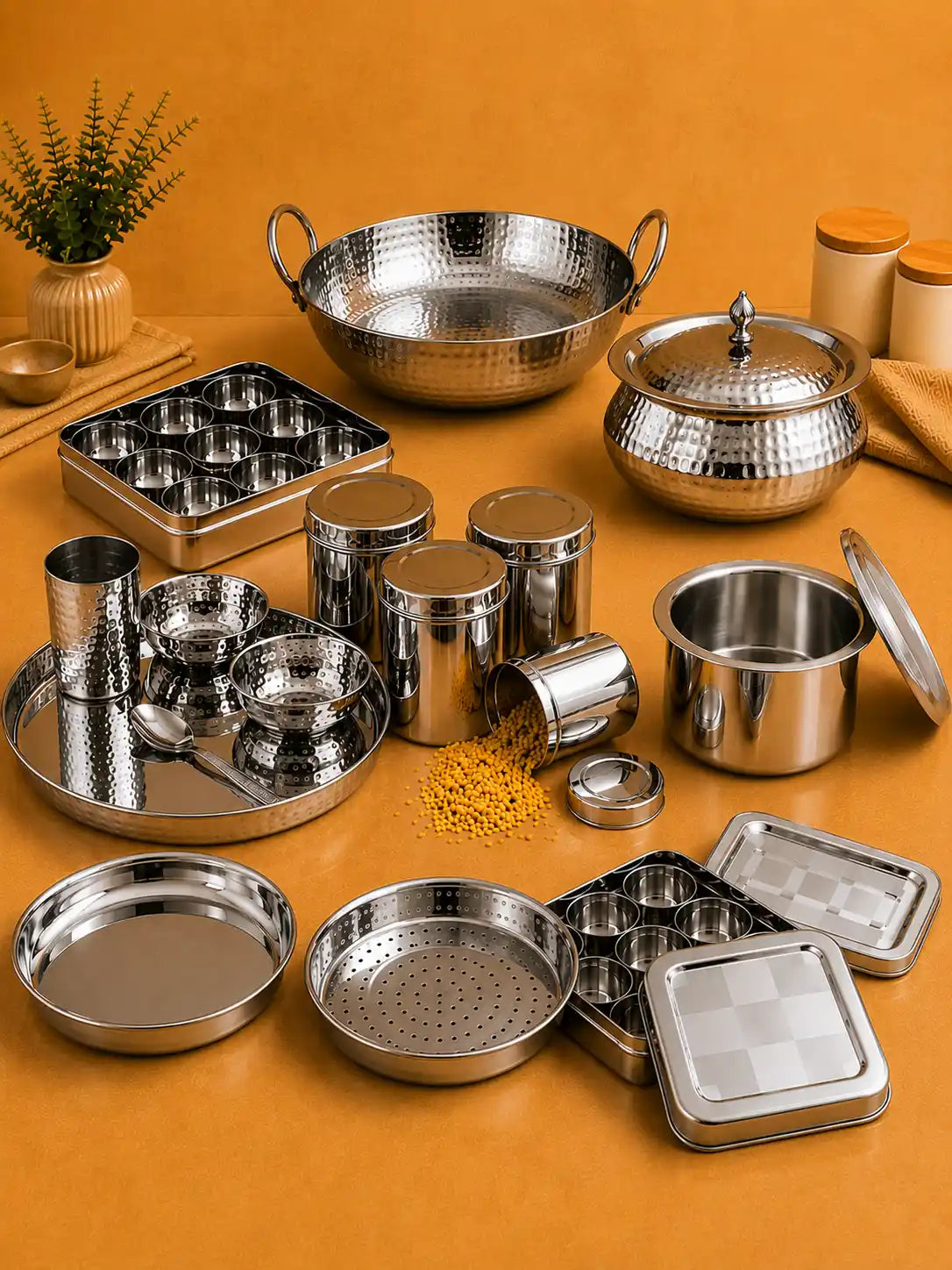 Premium stainless steel kitchenware collection featuring a hammered kadai, steamer plate, storage canisters, masala dabba, serving tray, and steel containers on a warm kitchen-themed background.