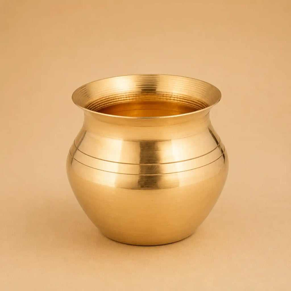A polished brass pot with a rounded body and flared rim, featuring horizontal engraved lines, set against a solid beige background.