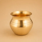 A polished brass pot with a rounded body and flared rim, featuring horizontal engraved lines, set against a solid beige background.