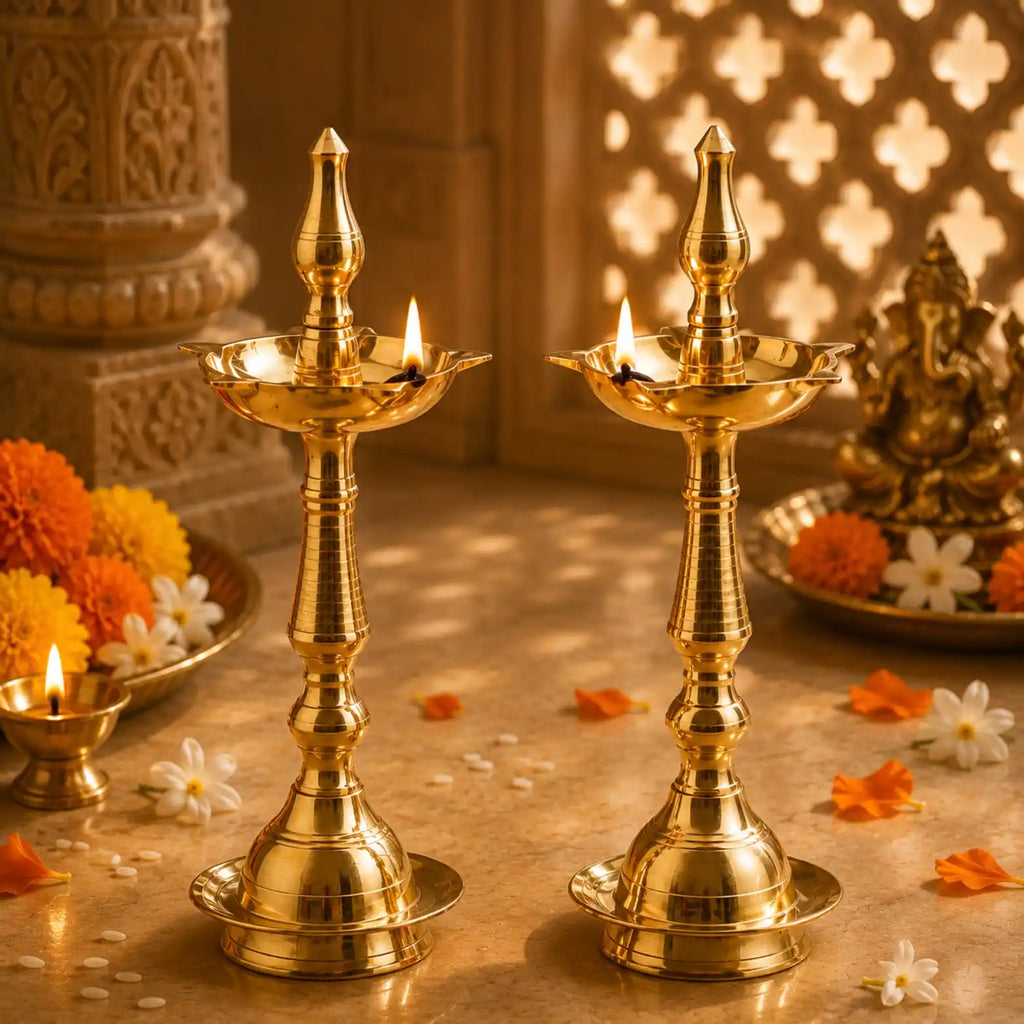 Two ornate brass oil lamps with lit wicks are displayed on a stone surface, surrounded by marigold and white flowers, with a decorative statue and carved pillar in the background.
