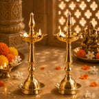 Two ornate brass oil lamps with lit wicks are displayed on a stone surface, surrounded by marigold and white flowers, with a decorative statue and carved pillar in the background.