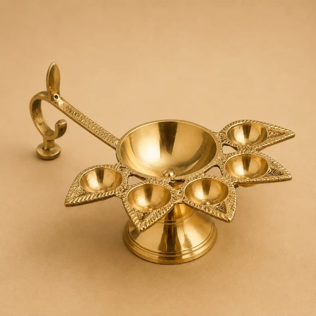 A polished brass five-light diya holder with an ornate curved handle, displayed on a solid beige background.