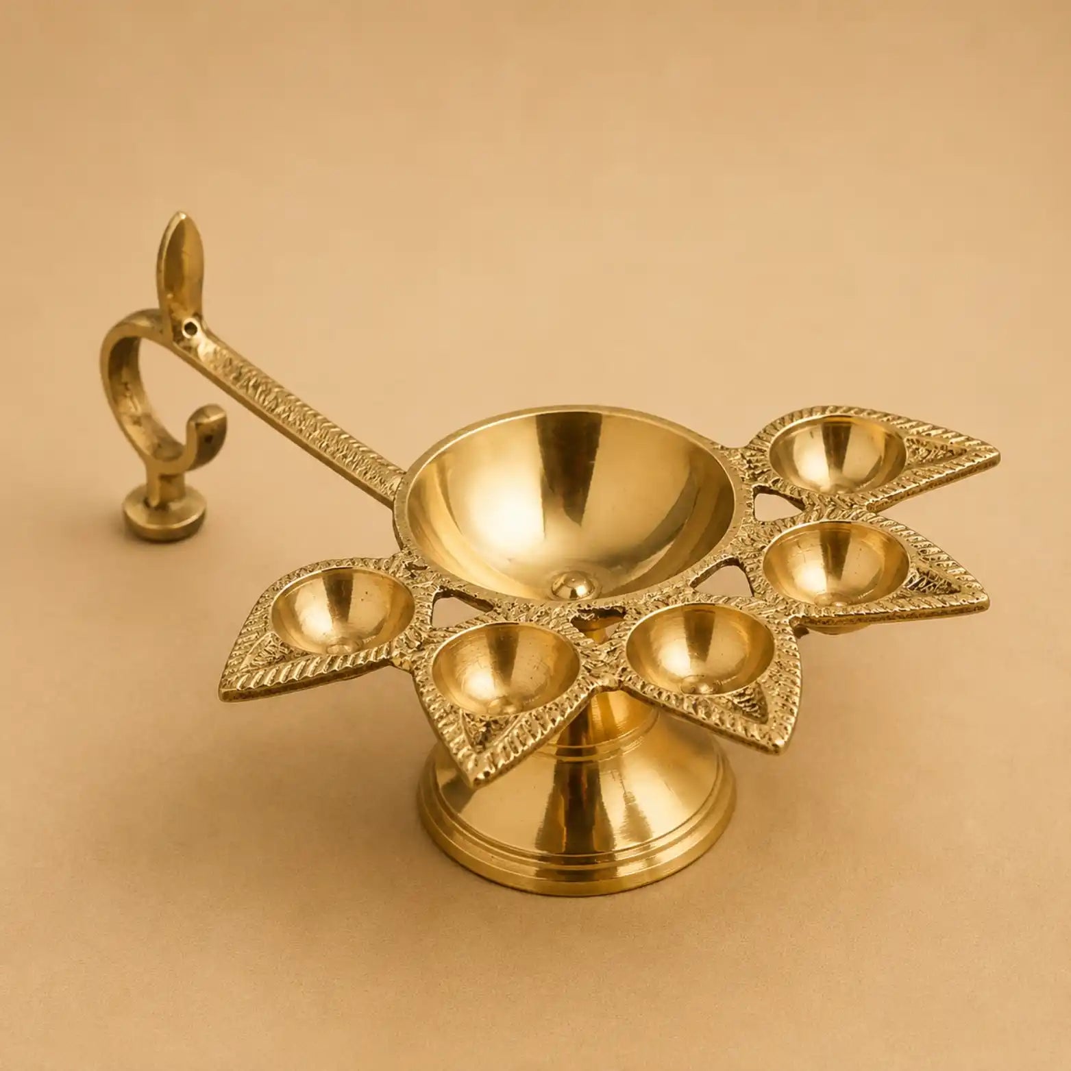 A polished brass five-light diya holder with an ornate curved handle, displayed on a solid beige background.