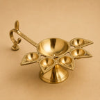 A polished brass five-light diya holder with an ornate curved handle, displayed on a solid beige background.