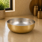 Gold metallic bowl on a beige surface with decorative jars and a plant in the background.