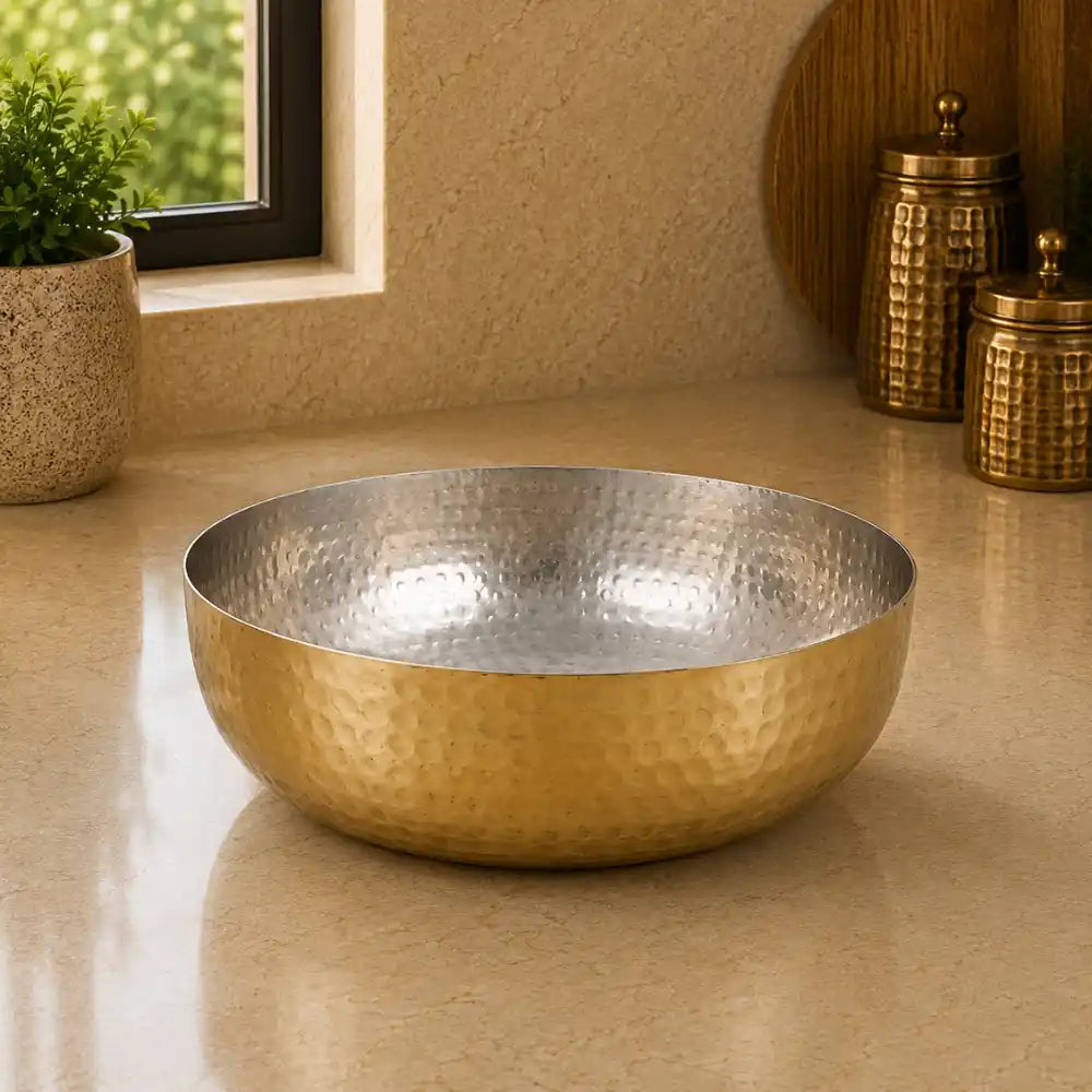 Gold metallic bowl on a beige surface with decorative jars and a plant in the background.