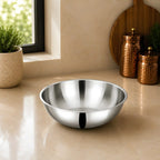 A shiny stainless steel mixing bowl placed on a beige countertop in a kitchen setting, with a window, potted plant, and copper canisters in the background.