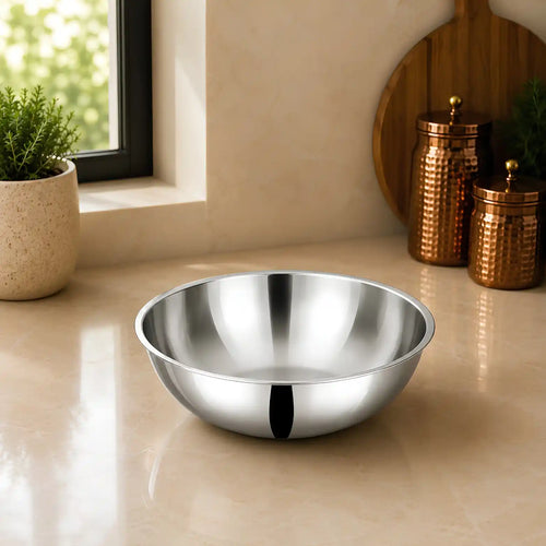 A shiny stainless steel mixing bowl placed on a beige countertop in a kitchen setting, with a window, potted plant, and copper canisters in the background.