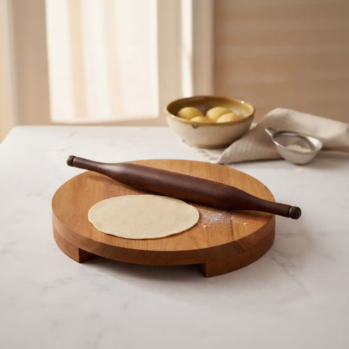 A round wooden rolling board with a dark wooden rolling pin and a round piece of dough, set on a light marble countertop with a bowl of dough balls in the background.