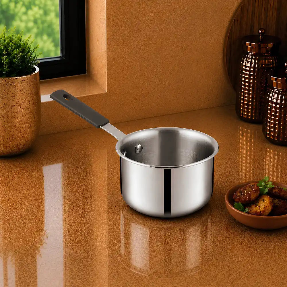 A stainless steel saucepan with a dark gray handle sits on a glossy brown countertop in a kitchen setting, with a window, potted plant, and decorative jars in the background.