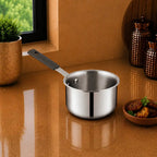 A stainless steel saucepan with a dark gray handle sits on a glossy brown countertop in a kitchen setting, with a window, potted plant, and decorative jars in the background.