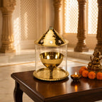 A brass oil lamp holder with a clear glass cylinder and conical lid, displayed on a dark wooden table in a warm, ornate indoor setting with a small lit diya and marigold garlands nearby.
