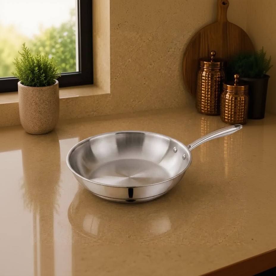 A stainless steel sauté pan with a long handle sits on a beige countertop in a kitchen setting, with a window, potted plant, and copper-colored canisters in the background.
