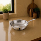 A stainless steel sauté pan with a long handle sits on a beige countertop in a kitchen setting, with a window, potted plant, and copper-colored canisters in the background.