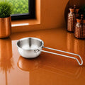 A small stainless steel saucepan with a long loop handle sits on a glossy copper-colored countertop in a kitchen setting, with a window, greenery, and decorative canisters in the background.
