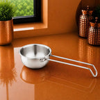 A small stainless steel saucepan with a long loop handle sits on a glossy copper-colored countertop in a kitchen setting, with a window, greenery, and decorative canisters in the background.