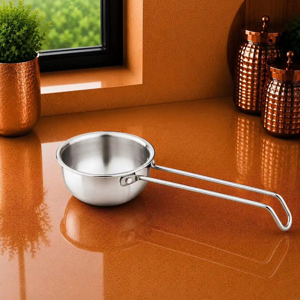 A small stainless steel saucepan with a long loop handle sits on a glossy copper-colored countertop in a kitchen setting, with a window, greenery, and decorative canisters in the background.