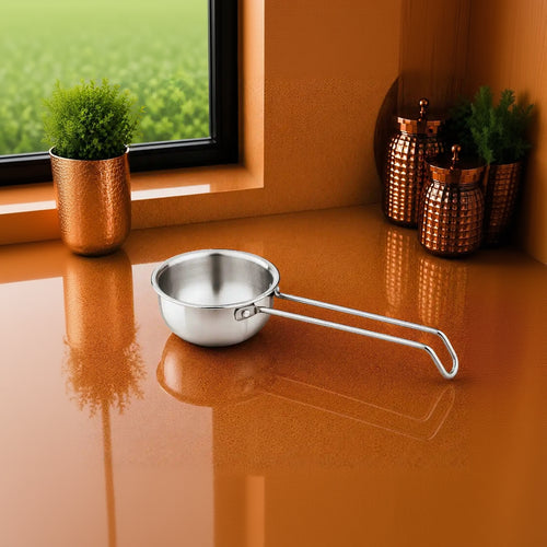 A small stainless steel saucepan with a long loop handle sits on a glossy copper-colored countertop in a kitchen setting, with a window, greenery, and decorative canisters in the background.