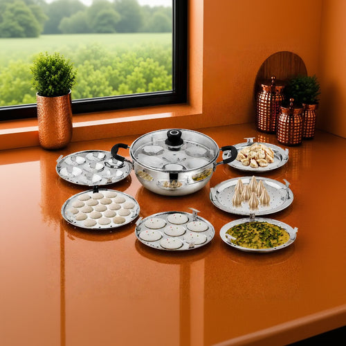 A stainless steel multi-tier steaming tray set displayed on an orange countertop near a window, with various dumplings and prepared foods arranged around the pots and pans.