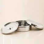 A stainless steel multi-compartment food container with its two lids off, displayed on a light beige background.