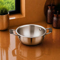 A stainless steel pot with two side handles sits on a glossy terracotta countertop in a kitchen setting, with a small plant and copper-colored canisters in the background.