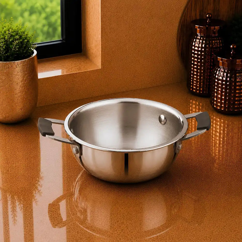 A stainless steel pot with two side handles sits on a glossy terracotta countertop in a kitchen setting, with a small plant and copper-colored canisters in the background.