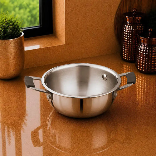 A stainless steel pot with two side handles sits on a glossy terracotta countertop in a kitchen setting, with a small plant and copper-colored canisters in the background.