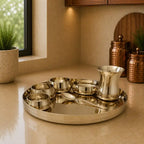 Set of silver platters, bowls, and a pitcher on a beige surface with a window and plants in the background.