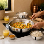 A stainless steel bowl filled with a colorful vegetable curry sits on a black induction cooktop in a kitchen setting. Hands are visible holding the bowl and spoon, with bowls of rice, flour, turmeric, and yellow ghee arranged around it.