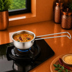 A stainless steel saucepan with a long handle sits on a black gas stove burner, cooking food on a warm brown countertop in a kitchen setting.