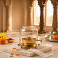 A brass oil lamp with a clear glass dome cover sits on a marble tray in a softly lit indoor setting with decorative pillars and marigold flowers.