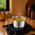 A stainless steel saucepan with a black handle sits on a black gas stovetop burner, with blue flame visible beneath. Steam rises from the soup inside, set in a warm kitchen scene with copper canisters, a window, and a small plant in the background.
