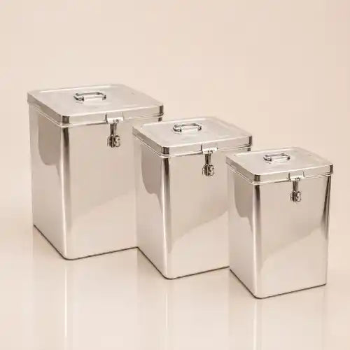 Three rectangular stainless steel canisters with lids and small front clasp closures, arranged by size on a warm beige studio background.