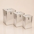 Three rectangular stainless steel canisters with lids and small front clasp closures, arranged by size on a warm beige studio background.
