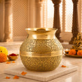 An ornate gold-toned metal pot with intricate floral engravings, placed on a white marble slab in a warm, softly lit indoor setting with decorative marigolds and a blurred background.