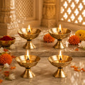 Four brass oil lamps (diyas) with lit wicks are arranged on a marble surface in a warm, decorated setting with marigolds and traditional architectural details in the background.