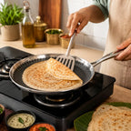A person wearing a beige apron uses a white spatula to flip a golden-brown dosa in a stainless steel pan on a black portable stove. Small bowls of chutney and a bottle of oil are visible in the background kitchen setting.