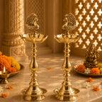 Two ornate brass oil lamps with peacock finials on a marble surface, surrounded by marigold flowers and a decorative background with warm lighting.