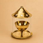 A polished gold-toned metal dreidel set inside a clear cylindrical glass or acrylic display case with a matching lid featuring star cutouts, photographed on a warm beige background.
