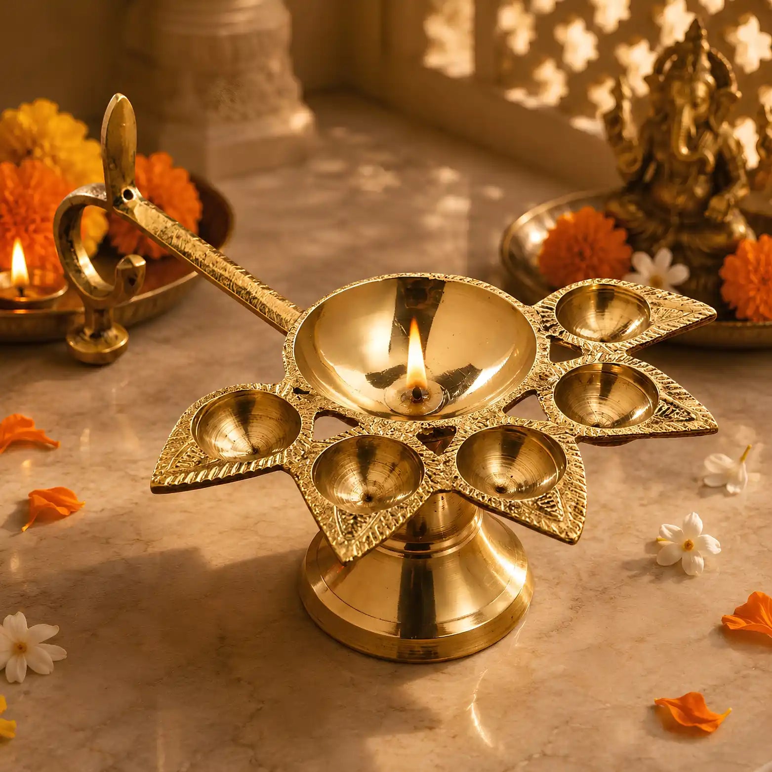 A brass diya lamp with five candle holders and a curved handle, lit with a flame, set on a marble surface with marigold flowers and decorative objects in the background.