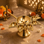 A brass diya lamp with five candle holders and a curved handle, lit with a flame, set on a marble surface with marigold flowers and decorative objects in the background.