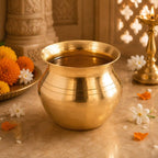 A polished brass pot with horizontal ridges, filled with water, set on a marble surface with marigolds, white flowers, and a brass candle holder in a warm, decorative setting.
