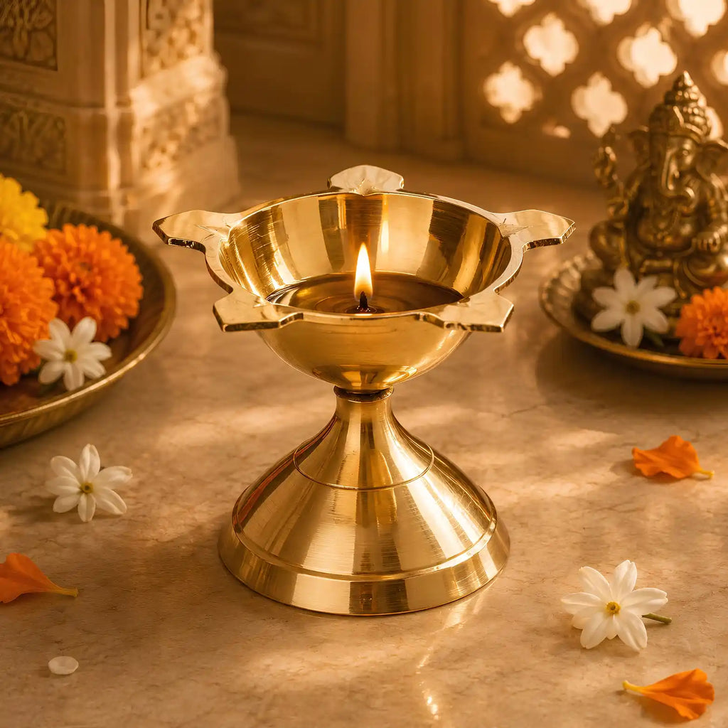 A polished brass oil lamp (diya) with a lit flame, set on a stone surface surrounded by marigold flowers, petals, and a decorative Ganesha idol in warm, golden light.