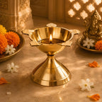 A polished brass oil lamp (diya) with a lit flame, set on a stone surface surrounded by marigold flowers, petals, and a decorative Ganesha idol in warm, golden light.