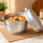 A stainless steel round baking pan with a matching lid leaning against it, filled with a baked pizza. The pan sits on a woven placemat on a light marble surface, with a potted plant and wooden cutting board blurred in the background.