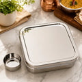 A square stainless steel bento lunch box with a matching lid, shown on a marble countertop with a small detached metal cup beside it. A potted plant, folded cloth, copper vessel, and bowl of food are visible in the background as styled kitchen props.