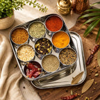 A stainless steel spice box set with multiple small round bowls filled with assorted whole and ground spices, arranged on a wooden surface with a matching tray, dried red chilies, bay leaves, and small plants.