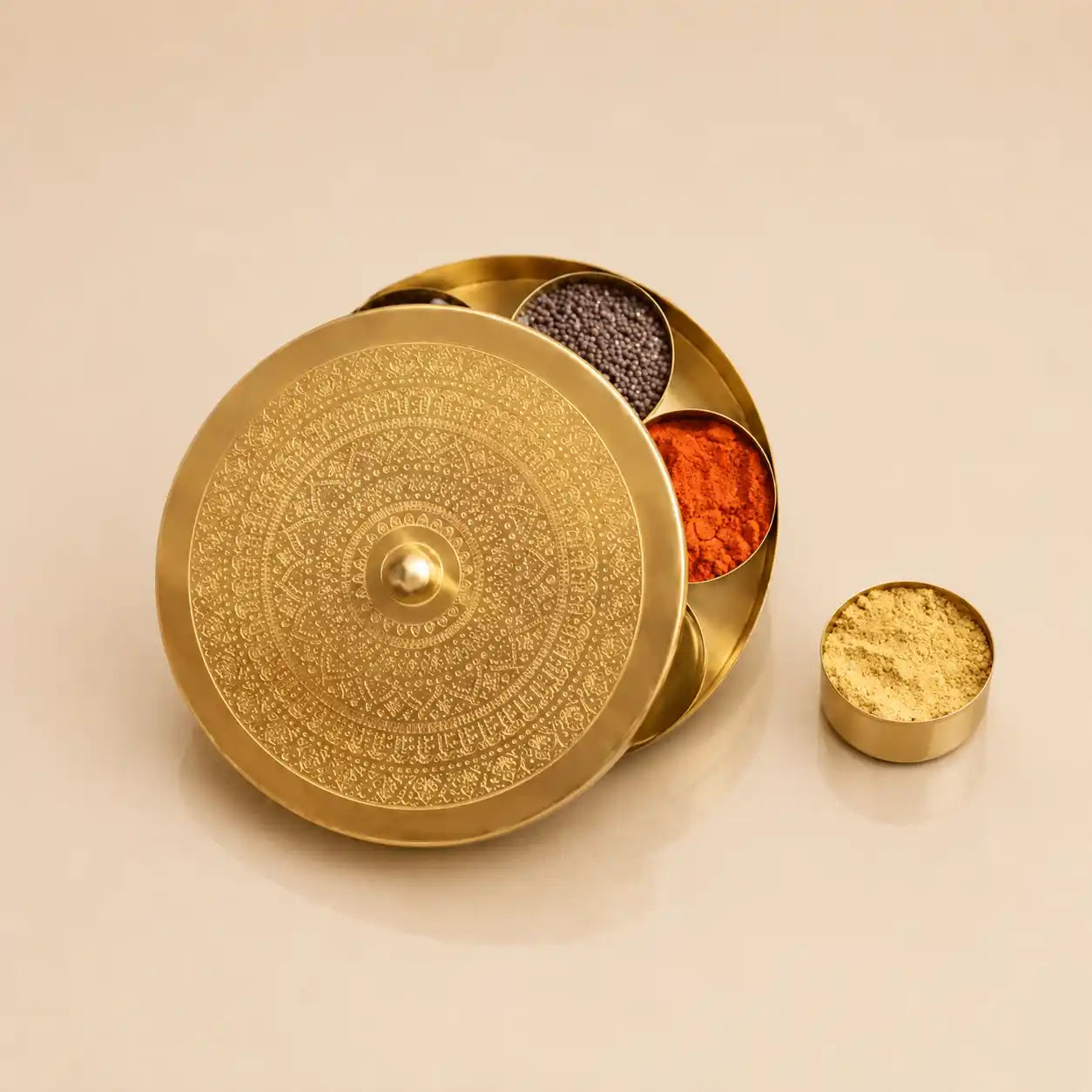 An ornate round brass spice container with a decorative embossed lid, shown open with several compartments containing colorful spices. A small separate vessel filled with golden powder sits beside it on a warm beige background.