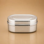 A stainless steel oval lunch box with a secure lid, shown on a warm beige background.