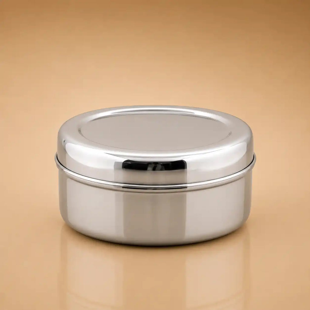 A round stainless steel lidded container with a polished metallic finish, shown on a warm beige gradient background.